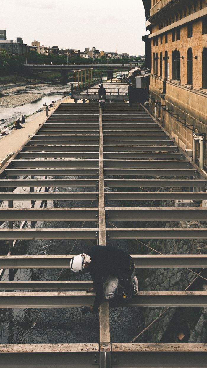 A construction worker carefully balances on steel beams by a riverside urban setting, showcasing industrial work.
